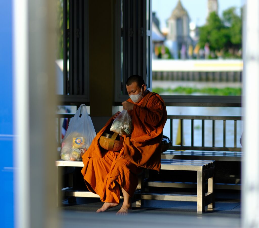 A Buddhist monk in orange robes with his alms bowl and morning offerings at Tha Tien pier, the Chao Phraya river behind