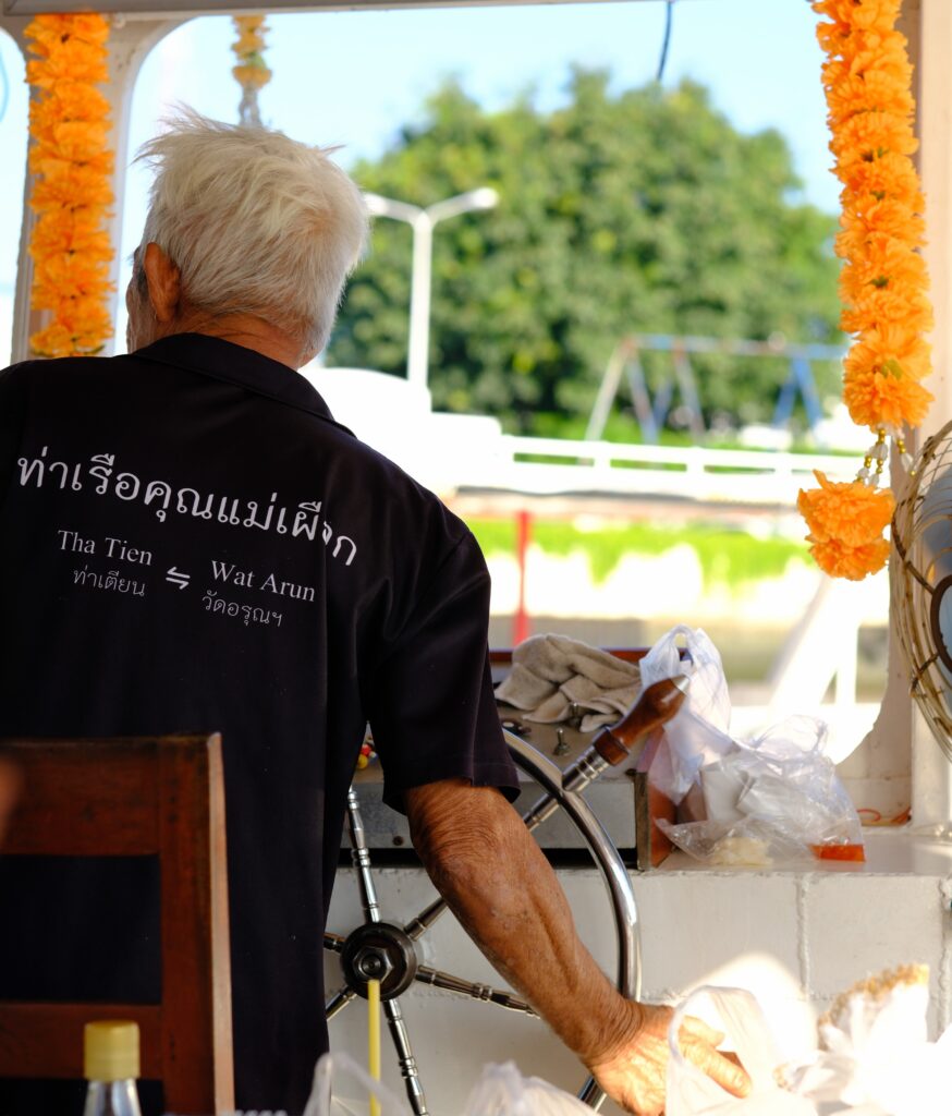 The ferryman at the wheel of the Tha Tien to Wat Arun boat, his shirt printed with the route in Thai and English