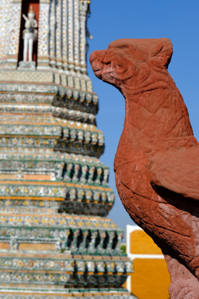 A weathered terracotta guardian figure in profile against the prang of Wat Arun