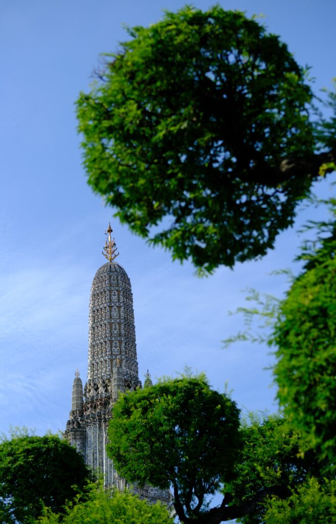 The central prang of Wat Arun, a model of Mount Meru, rising above the trees against a blue sky